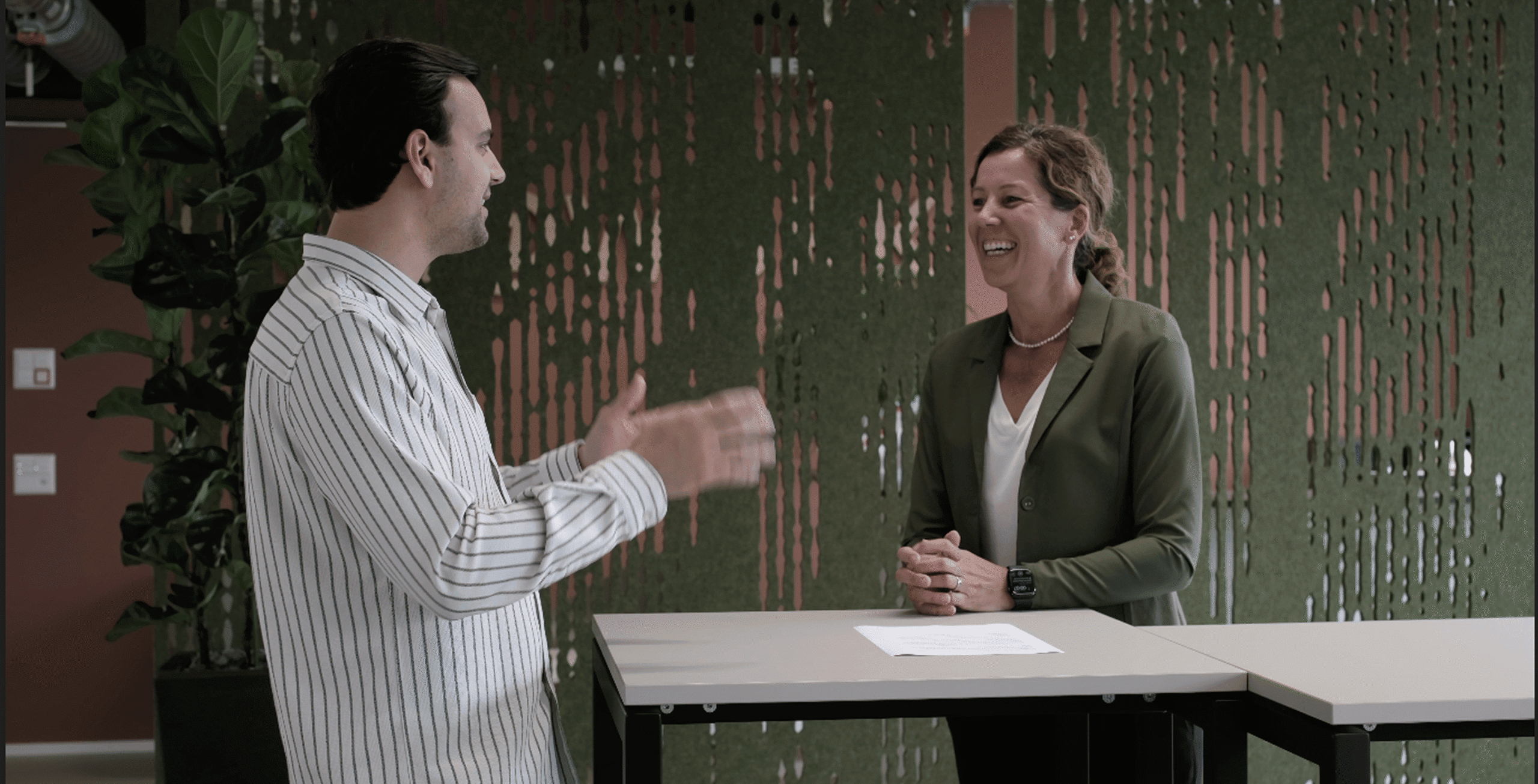 Zwei Personen stehen an einem hohen Tisch und unterhalten sich. Eine gestikuliert dabei, die andere lächelt. Im Hintergrund sind eine grüne Dekoplatte und eine Topfpflanze zu sehen.