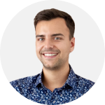 Lächelnder Mann mit kurzen dunklen Haaren, der ein blau gemustertes Hemd trägt, abgebildet vor einem schlichten hellen Hintergrund.