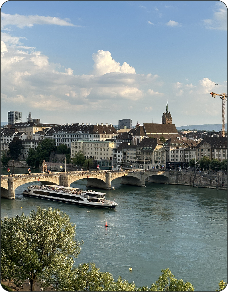 Ein Boot fährt unter einer steinernen Brücke über einen Fluss hindurch. Im Hintergrund sind historische Gebäude und ein Kirchturm zu sehen, während der Himmel teilweise bewölkt ist.